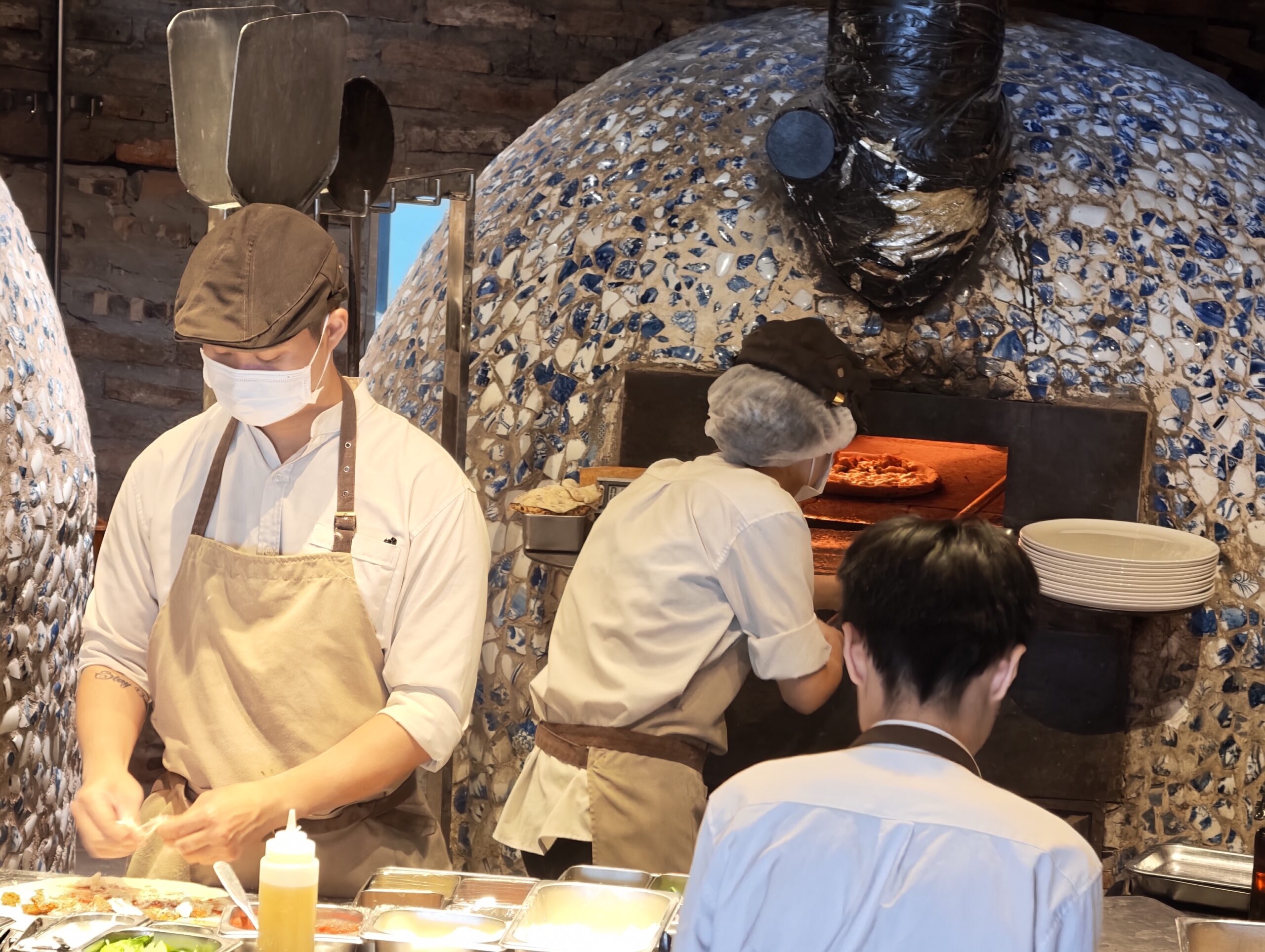 帶著口罩與帽子的 Pizza 4P's 廚師，在開放式吧台區專心備料與製作披薩，後方是吸睛的巨大馬賽克窯爐。